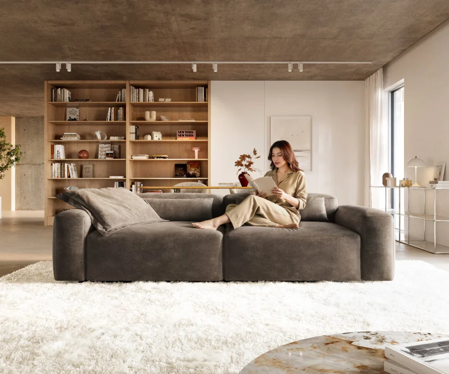 Canapé Freestyle en velours gris taupe, une femme lit assise dessus devant une bibliothèque.