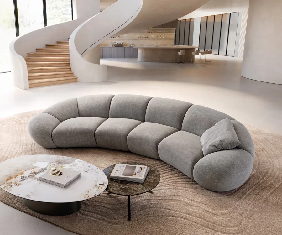Canapé gris courbé en tissu, vu de biais, avec coussin, tables basses et escalier au fond.